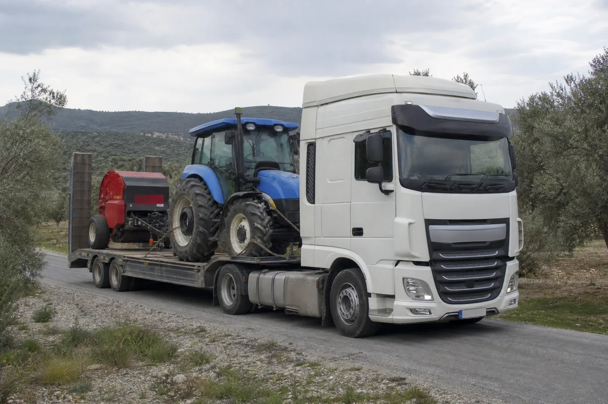 2 akslı lowbed dorse üzerinde zincirle bağlanmış mavi traktör ve balya makinesi, zeytinlikler arasında